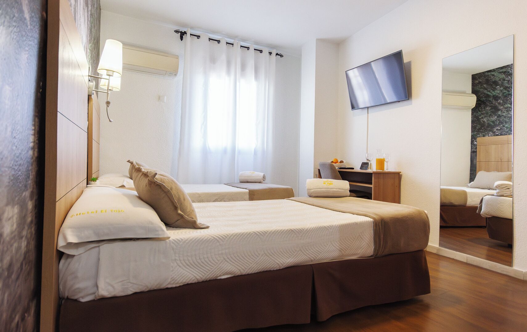 Ein Hotelzimmer mit zwei Betten, einem Flachbildfernseher, einem Spiegel, einem Schreibtisch mit Stuhl und einem fenster mit Vorhängen.