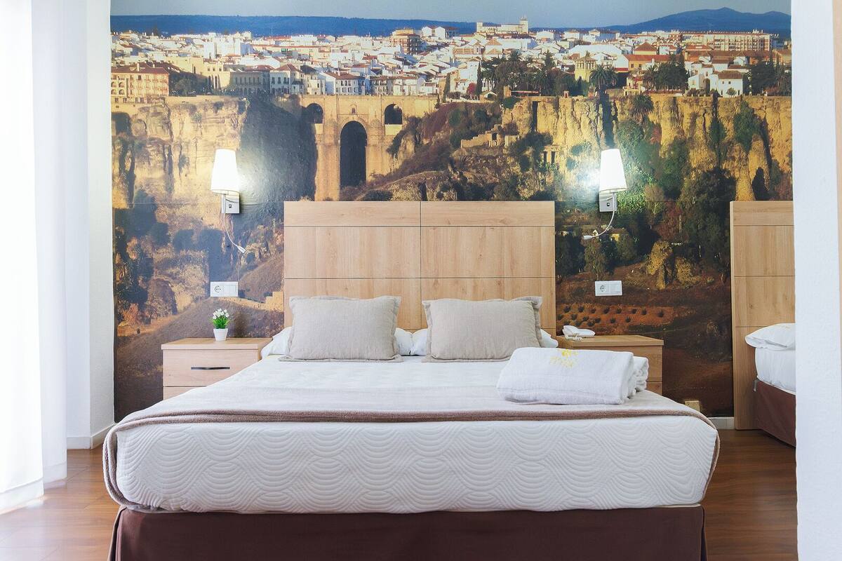 Ein Hotelzimmer mit Bett, Holz-Kopfteil, zwei Nachttischlampen, einem Nachttisch und einem großen Wandbild einer Stadtansicht.