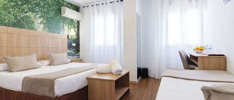 A hotel room with a bed, a desk with a chair, a mirror, and a view of greenery through the window.
