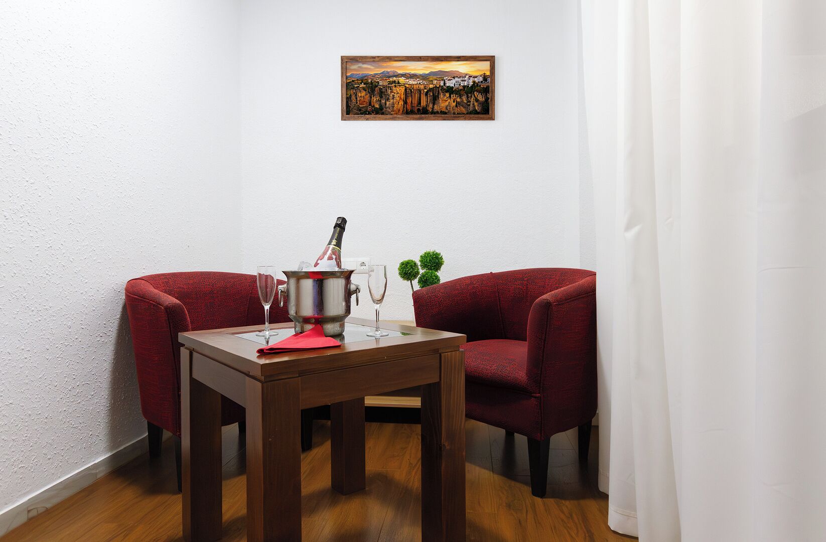 A room with a wooden table, two red armchairs, a painting on the wall, and a bottle of champagne.