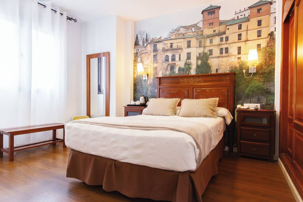 Ein Hotelzimmer mit einem großen Bett, einem Holzkopfstück, Nachttischen, einer Bank und einem Wandgemälde einer Stadtansicht.