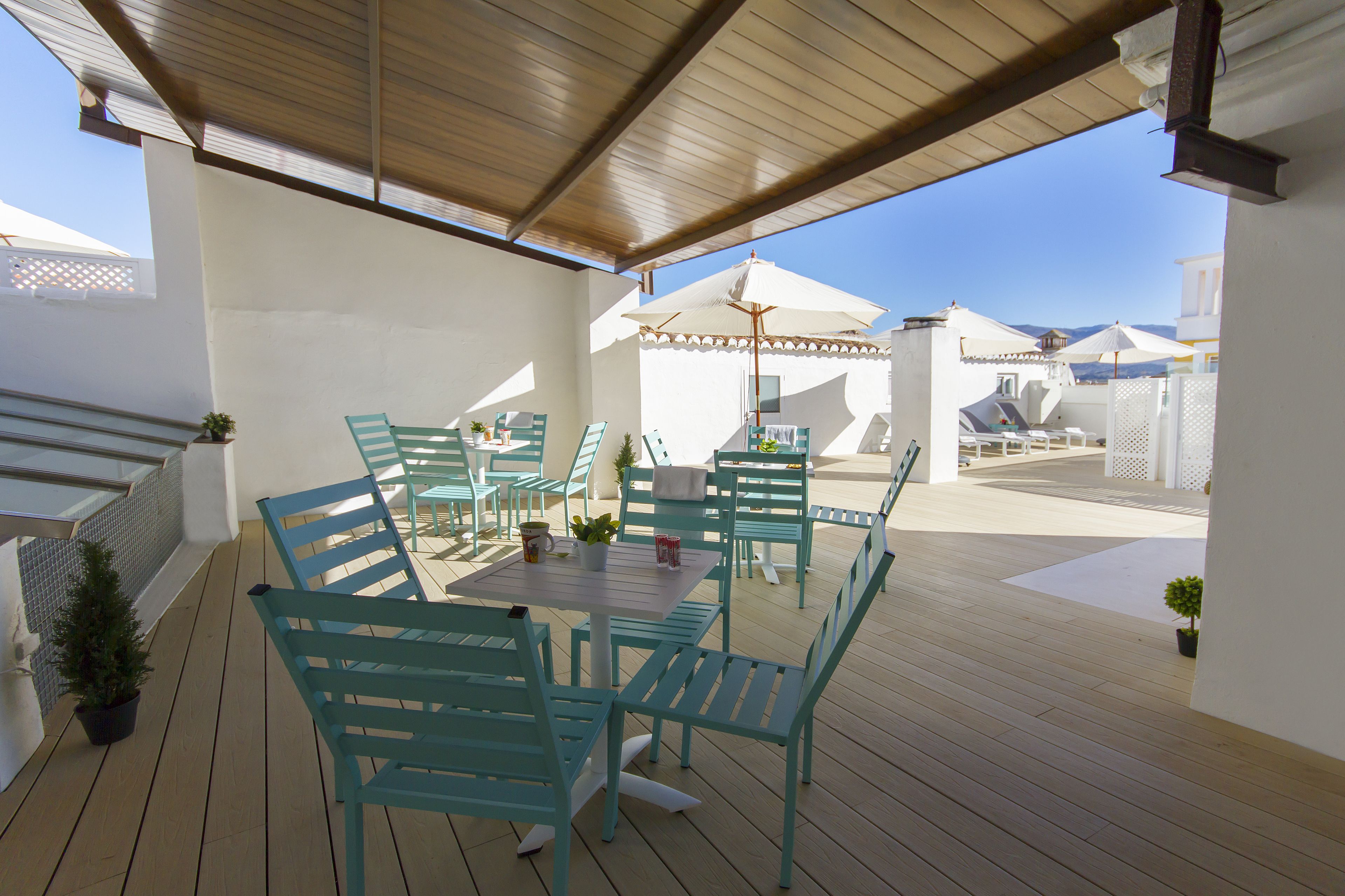 An outdoor patio with wooden flooring, turquoise chairs, and tables, featuring umbrellas and potted plants.