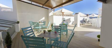 An outdoor patio with wooden flooring, turquoise chairs, and tables, featuring umbrellas and potted plants.