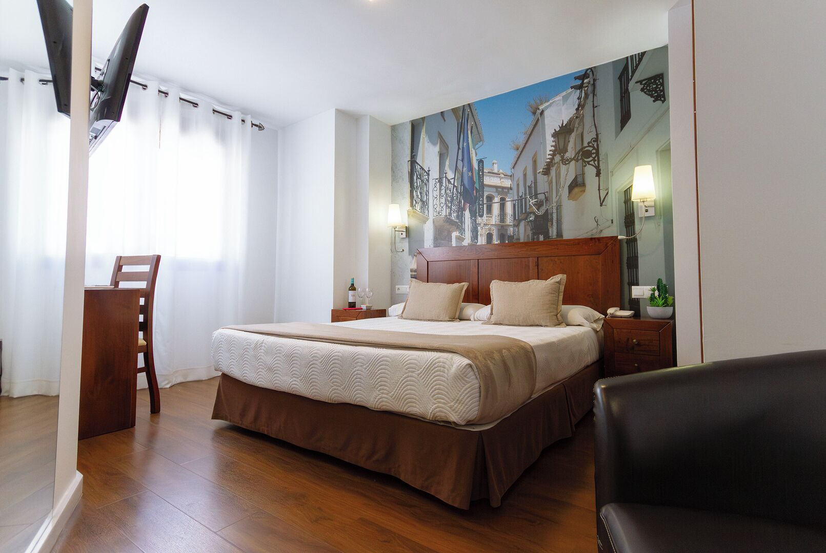 A hotel room with a bed, a desk, a chair, a painting of a street scene, and a lamp.
