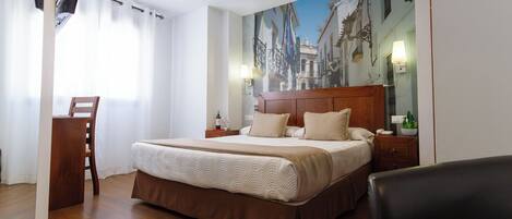 A hotel room with a bed, a desk, a chair, a painting of a street scene, and a lamp.