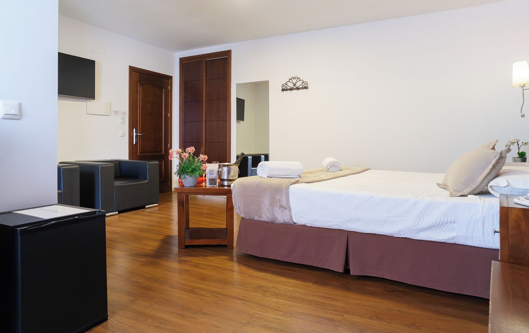 A hotel room with a bed, a desk, a chair, a small table with flowers, and a wooden floor.