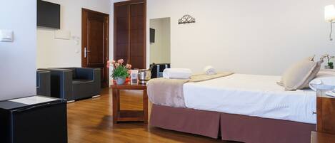 A hotel room with a bed, a desk, a chair, a small table with flowers, and a wooden floor.