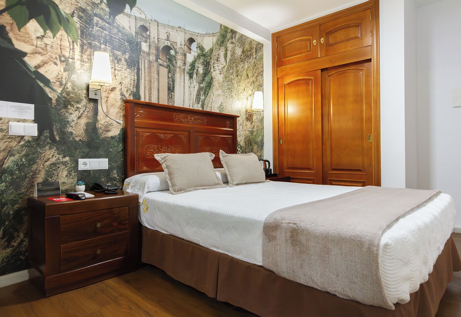 A hotel room with a bed, wooden wardrobe, nightstand, and a wall with a mural.
