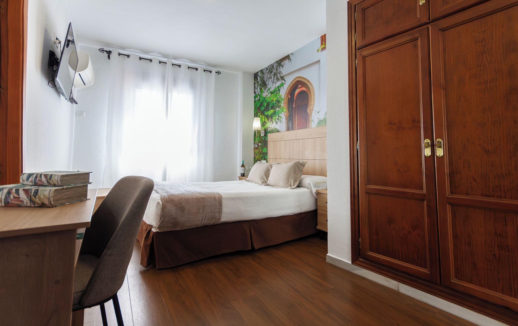 A bedroom with a bed, a desk with books, a chair, a wardrobe, and a wall-mounted lamp.
