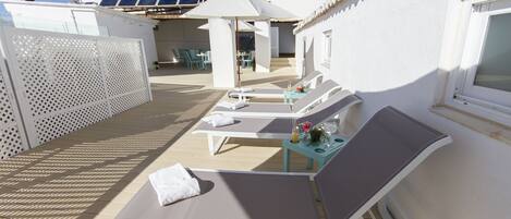 A sunlit outdoor lounge area with white lounge chairs, a table with a drink and a flower, and solar panels on the roof.