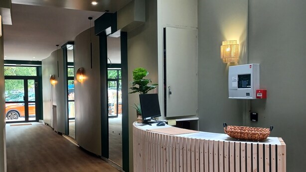 A reception area with a wooden reception desk, a potted plant, a wall-mounted security camera, and a fire alarm. - Hotel Le Colibri (Aix-les-Bains)