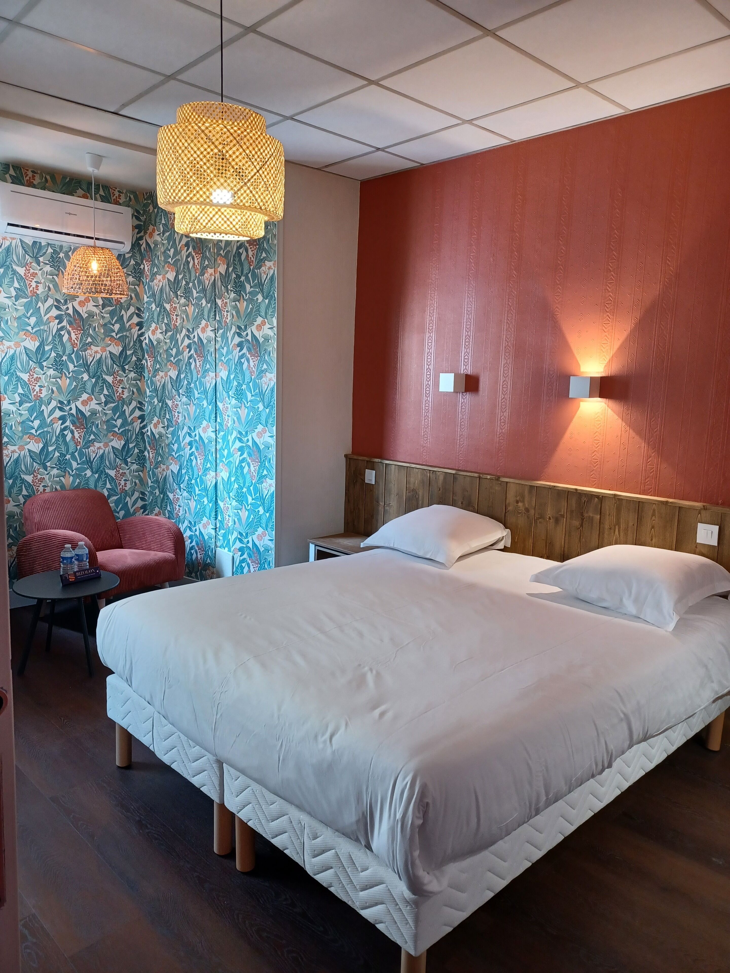 Una habitación de hotel con una cama grande, cabecera de madera, pared de acento roja, pared con papel tapiz floral, una silla roja y una lámpara colgante.