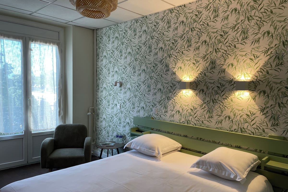 A hotel room with a bed, two pillows, a chair, a table with a bottle, and a wall with a bamboo pattern.