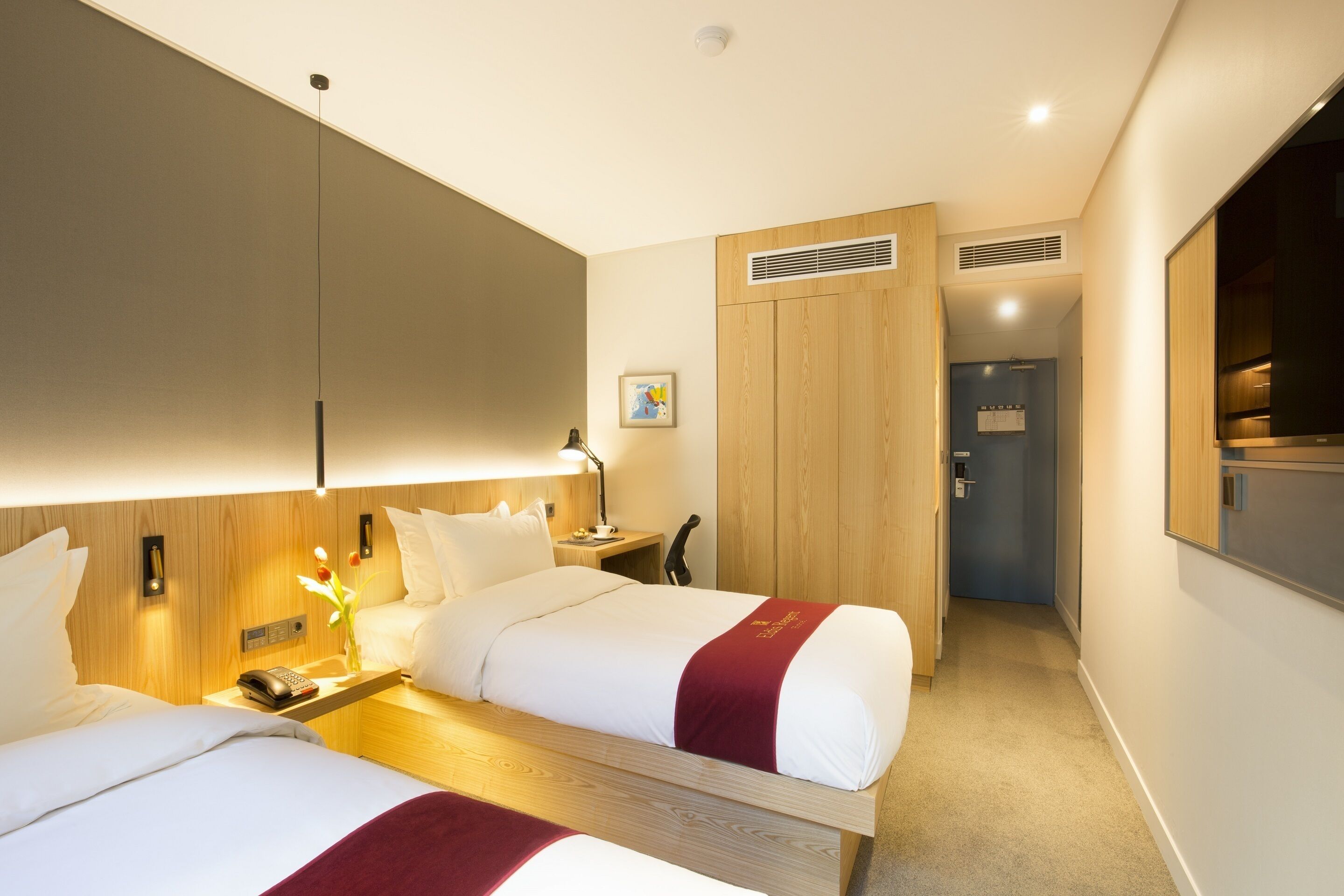 A hotel room with two beds, a wooden headboard, a bedside table with a lamp, a phone, and a picture on the wall.