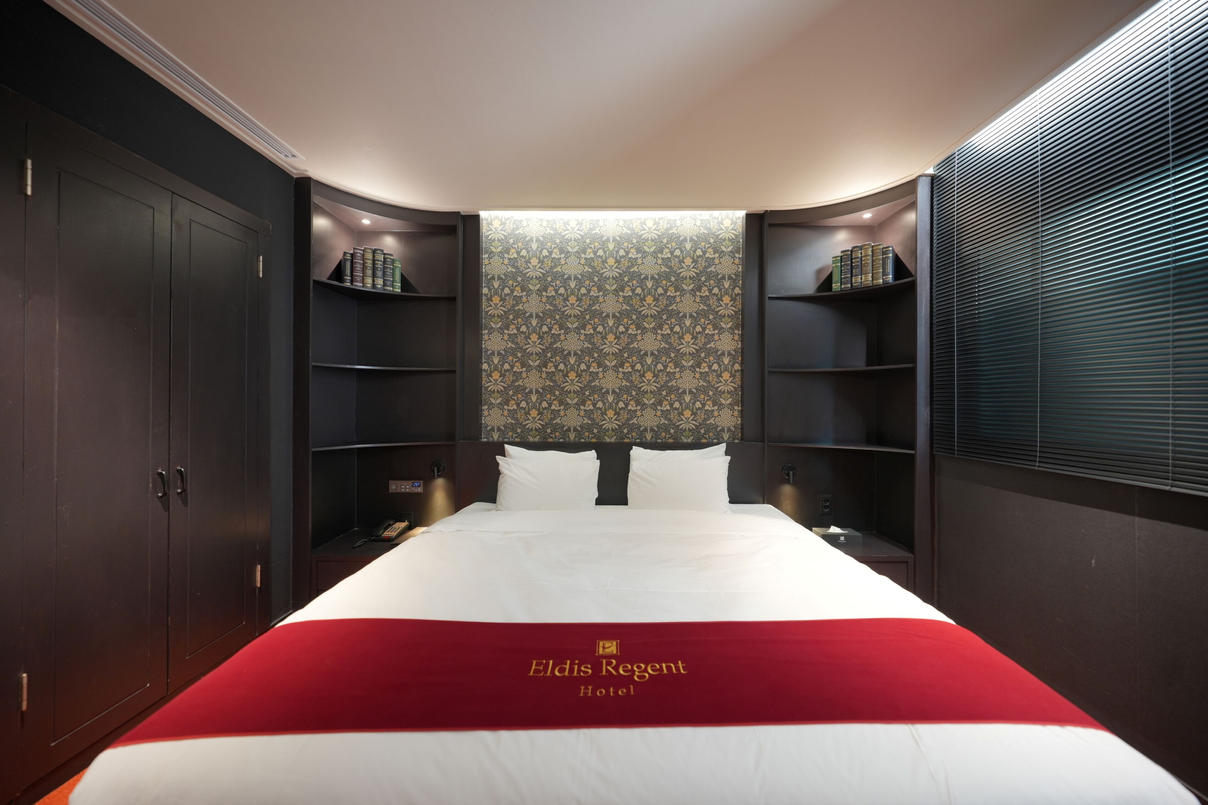 A hotel room with a large bed, dark wood wardrobe, and built-in shelving with books.