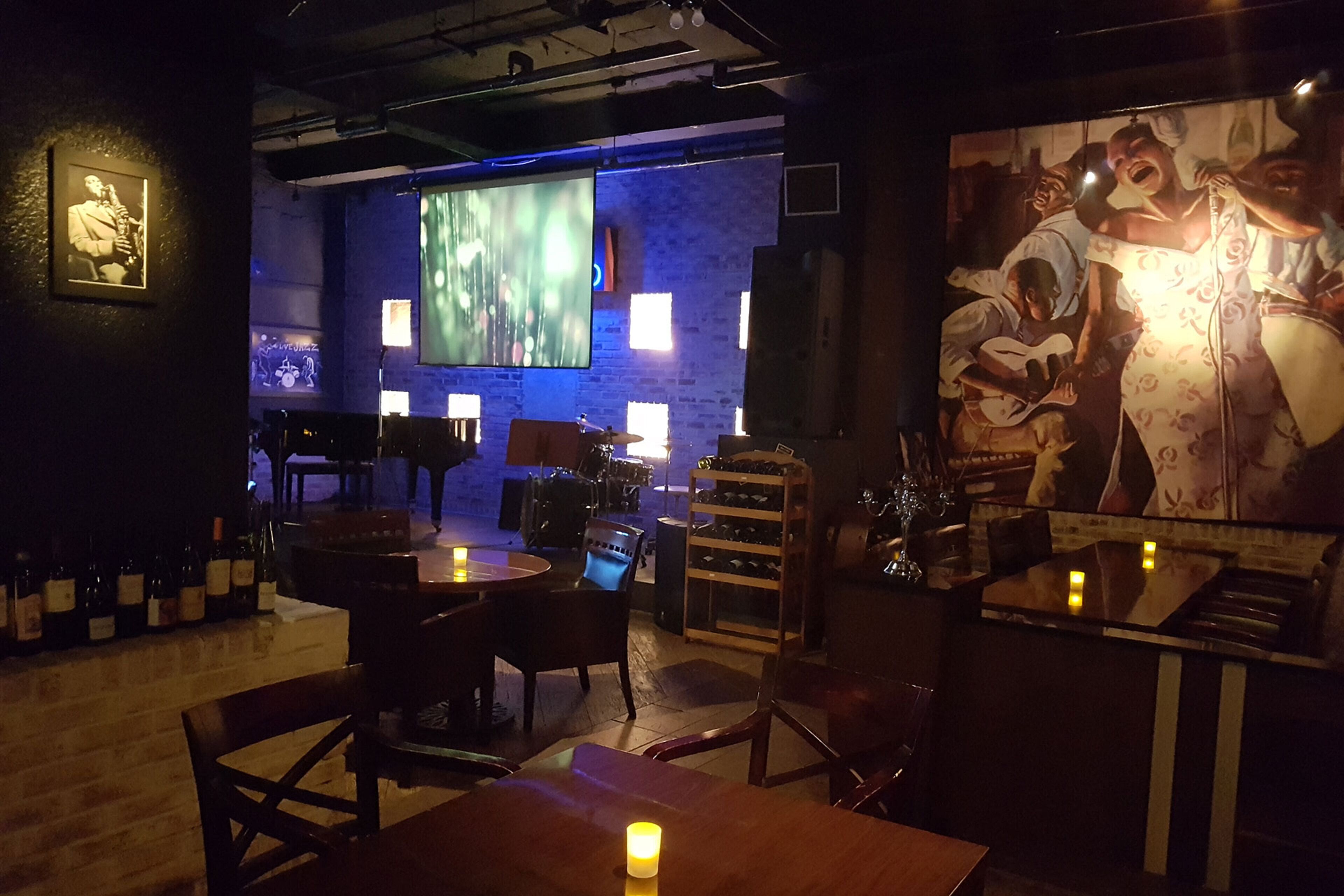 A dimly lit bar area with wooden tables and chairs, a large screen displaying a performance, and a mural on the wall.
