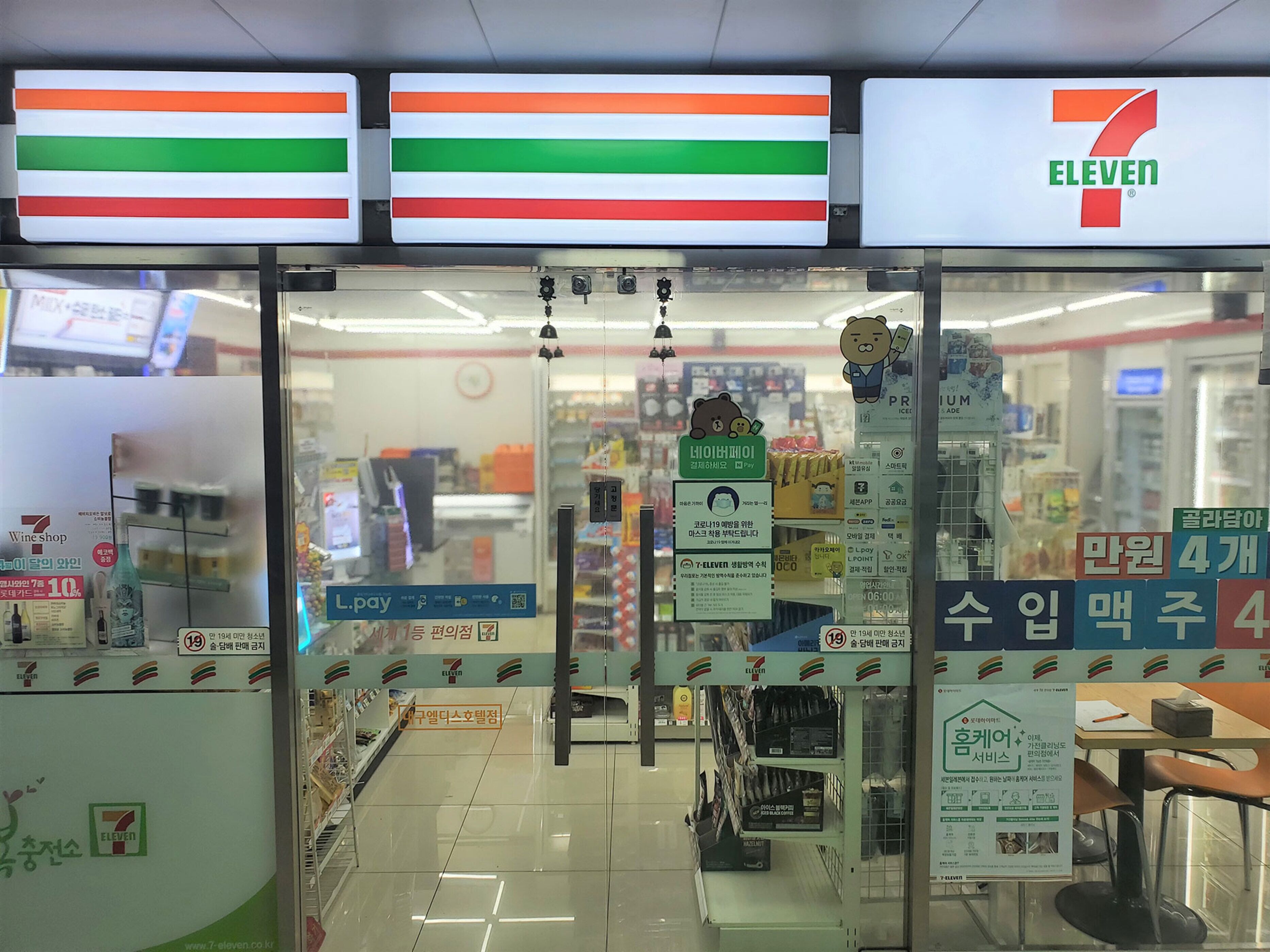 A 7-Eleven convenience store with a clear glass front, L.Pay payment options, and various product displays.