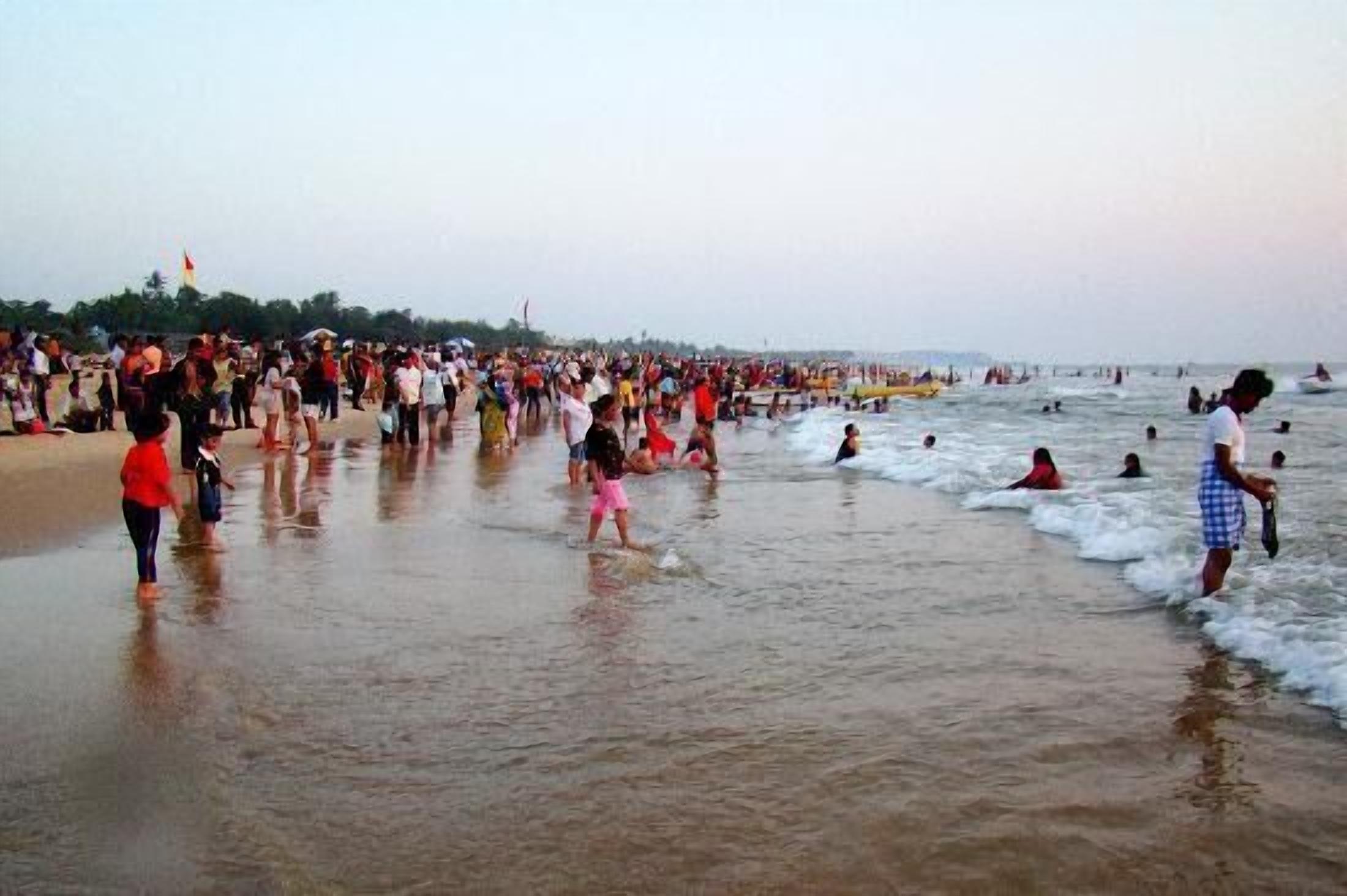 A crowded beach with people walking and swimming in the water.