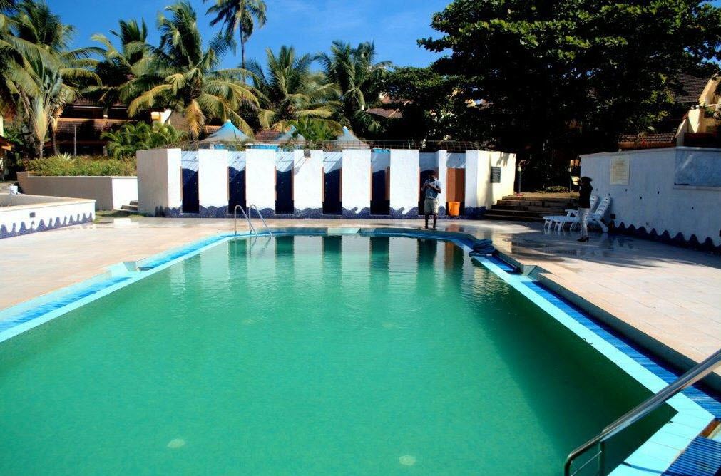 Una piscina con agua cristalina azul, rodeada por un edificio blanco con varias puertas y una zona cubierta.