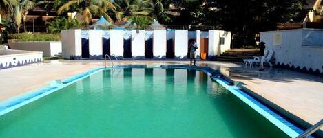 Una piscina con agua cristalina azul, rodeada por un edificio blanco con varias puertas y una zona cubierta.