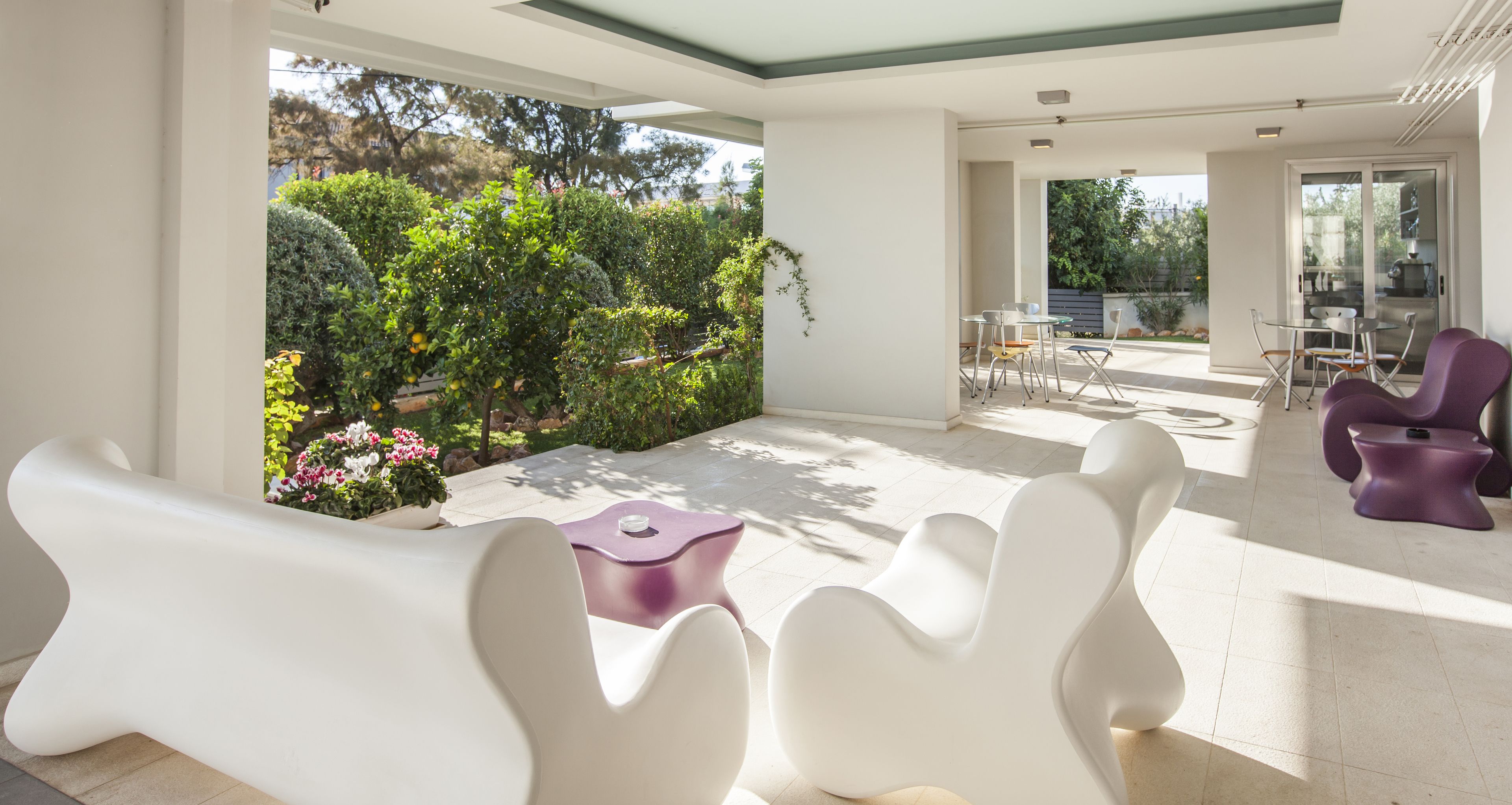 A modern outdoor patio with white and purple furniture, surrounded by lush greenery and a view of the city.