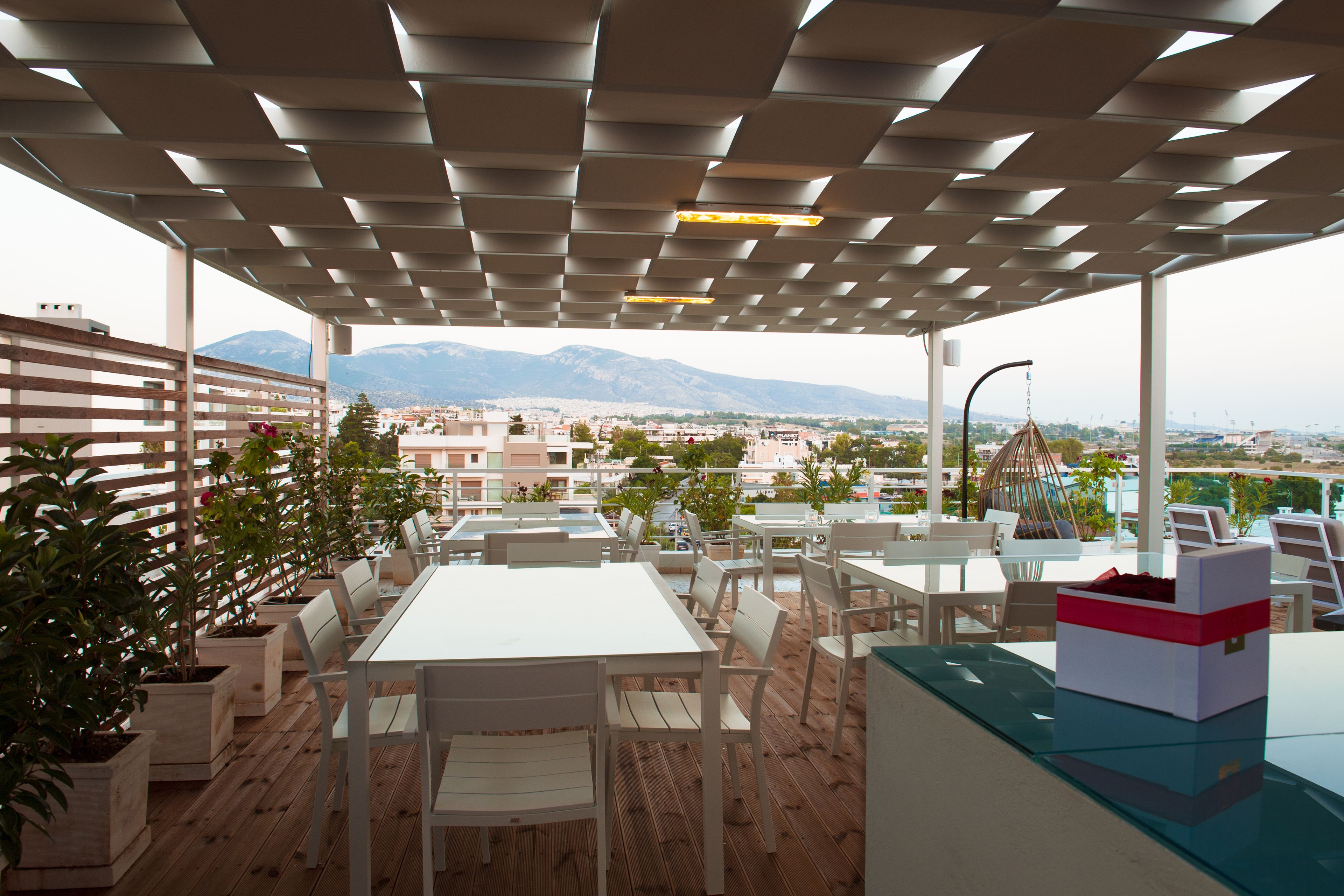 An outdoor terrace with white tables and chairs, potted plants, and a view of the city and mountains in the background.