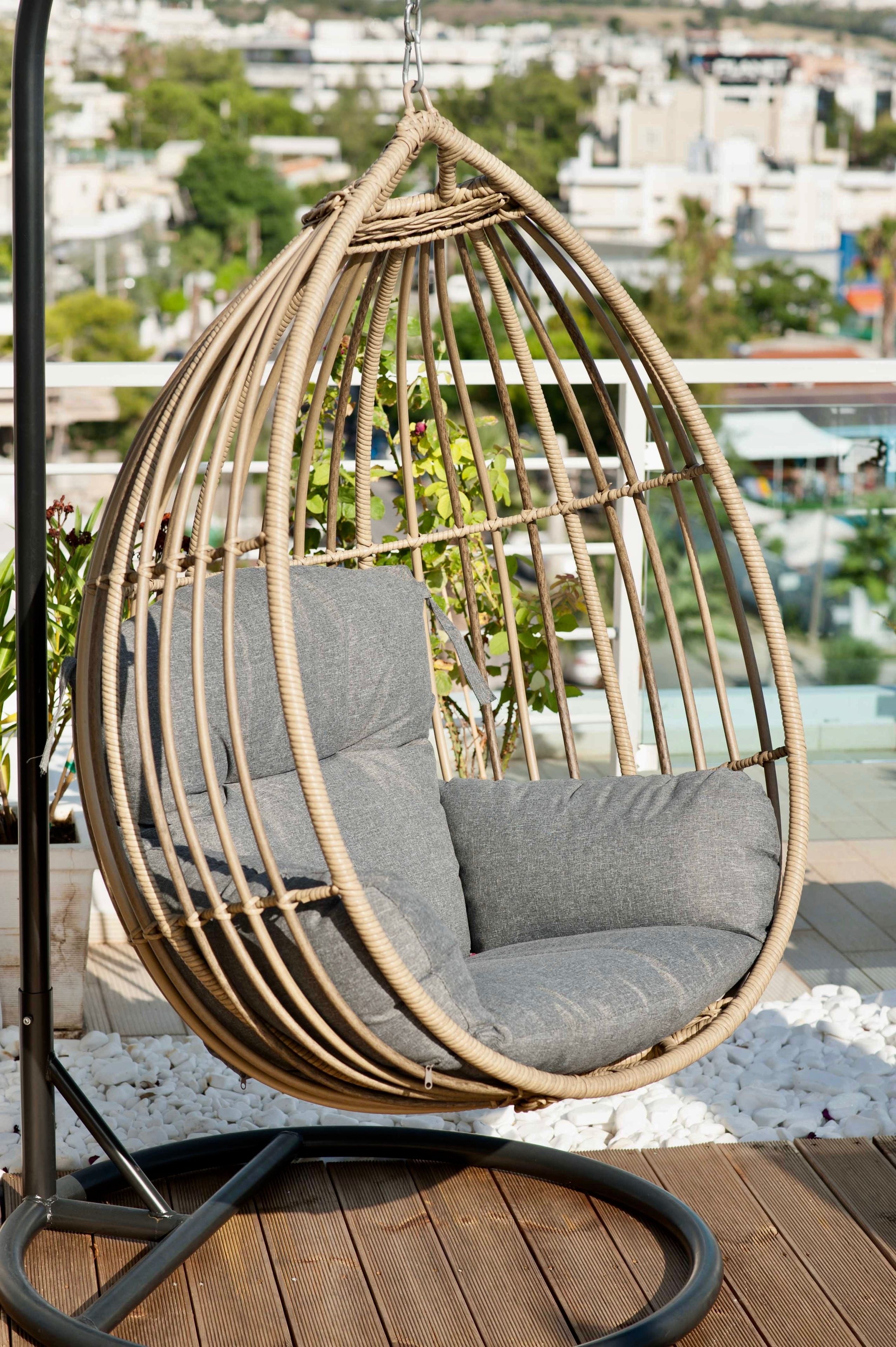 A wicker hanging chair with grey cushions on a wooden deck, overlooking a cityscape.