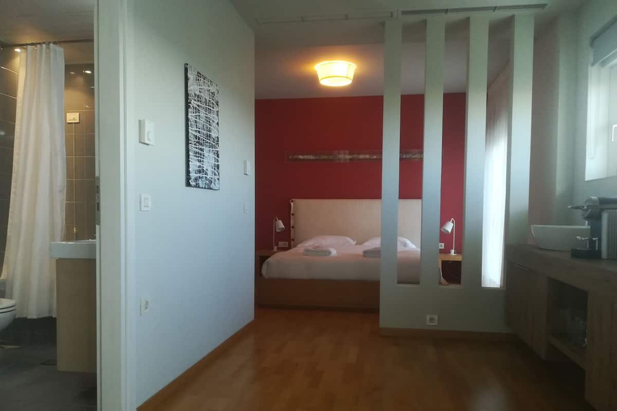 A hotel room with a wooden floor, a bed with white bedding, a red accent wall, a ceiling light, and a bathroom with a shower curtain.