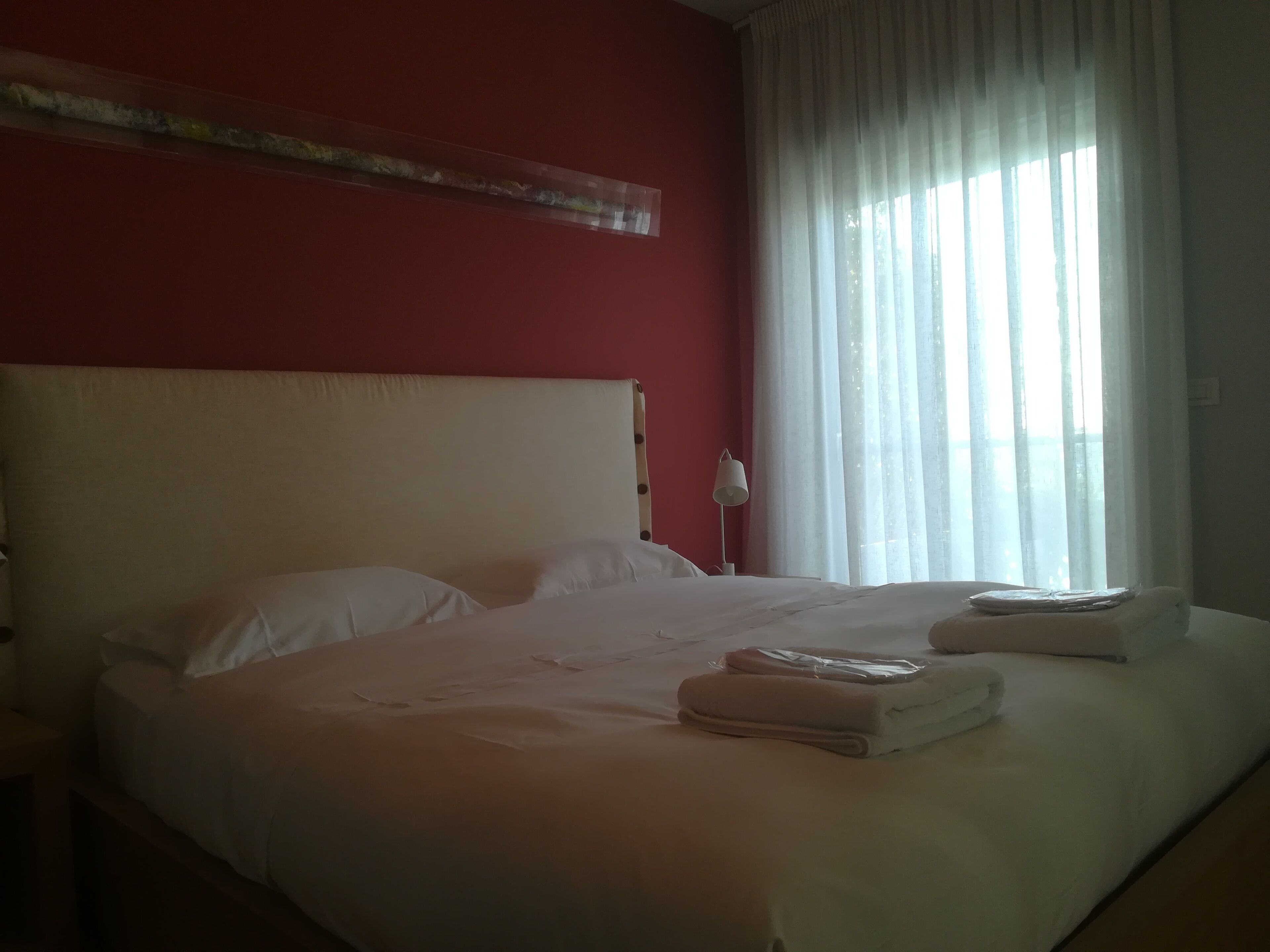 Ein Hotelzimmer mit einem großen Bett, weißer Bettwäsche, einer roten Wand und einem Fenster mit durchsichtigen Vorhängen.