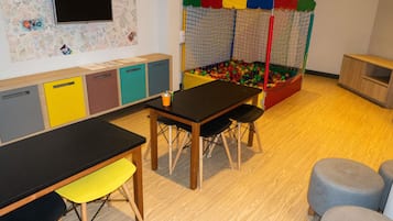 Un área de juegos con piscina de pelotas, una mesa con sillas y un televisor en la pared.