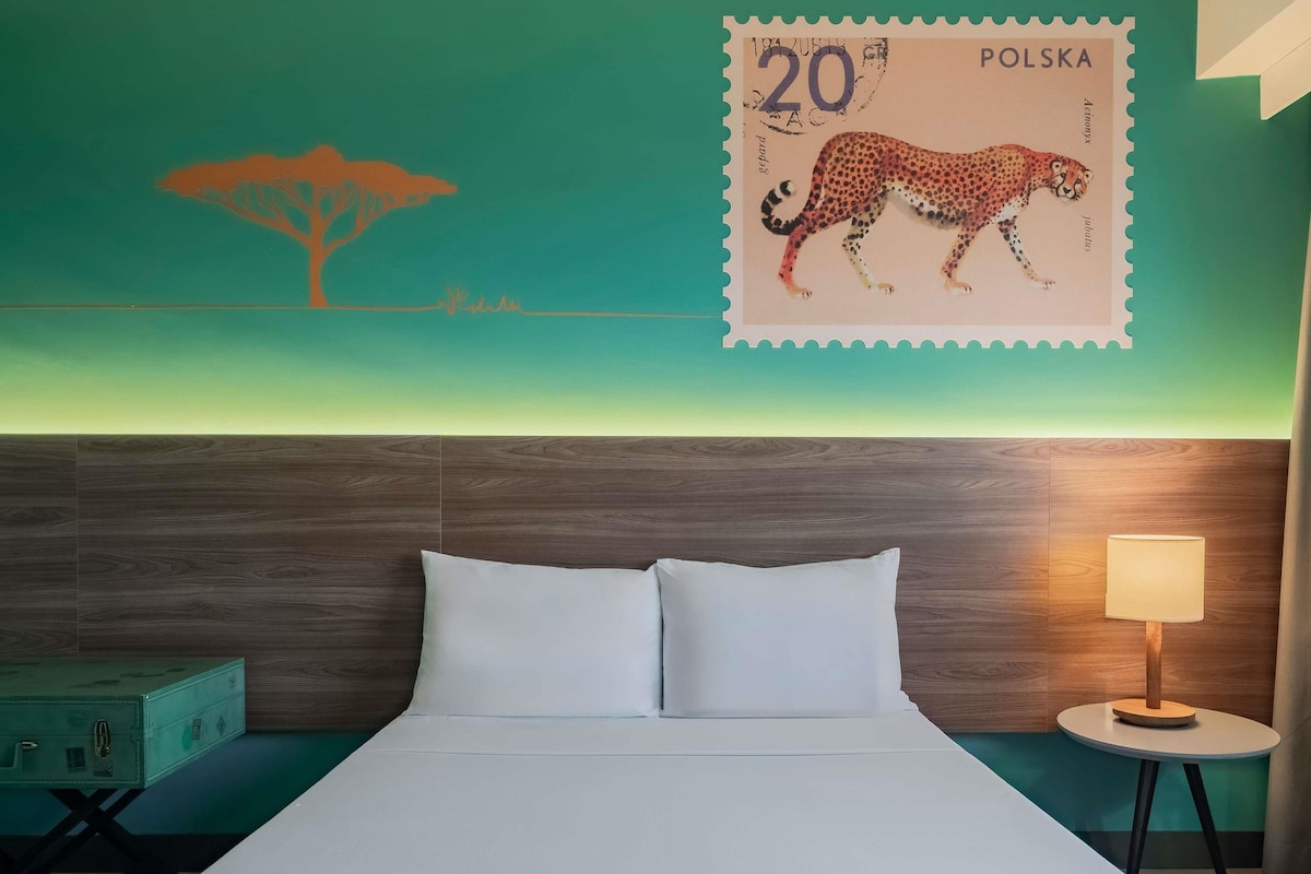 Una habitación de hotel con una cama, una mesita de noche, una lámpara, un cuadro colgado en la pared y un sello postal polaco.