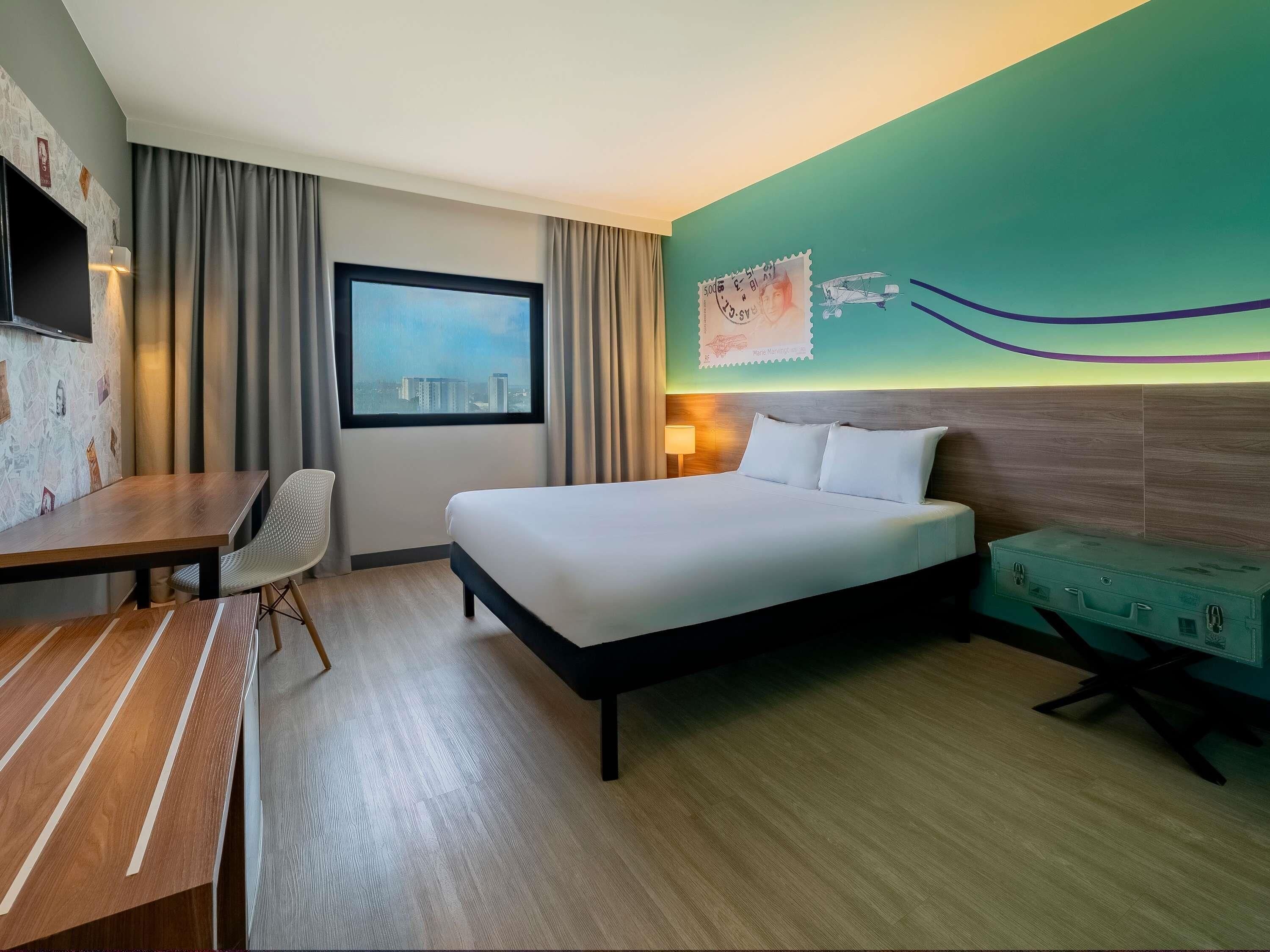 Une chambre d’hôtel avec un lit, un bureau, une chaise, une valise, une fenêtre donnant sur la ville et un mur orné d’une fresque.