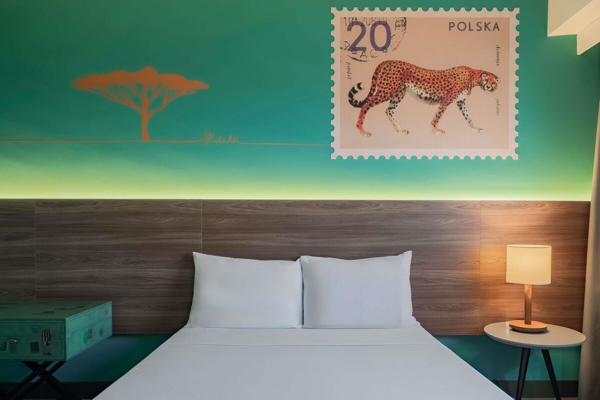 A hotel room with a bed, a nightstand, a lamp, a wall with a leopard stamp, and a teal-colored suitcase.