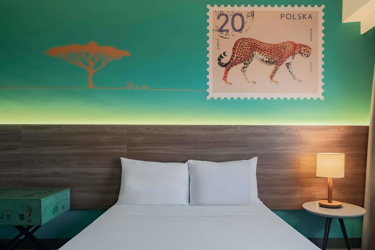 Une chambre d’hôtel avec un lit, une table de chevet, une lampe, un tableau accroché au mur et un timbre-poste polonais.