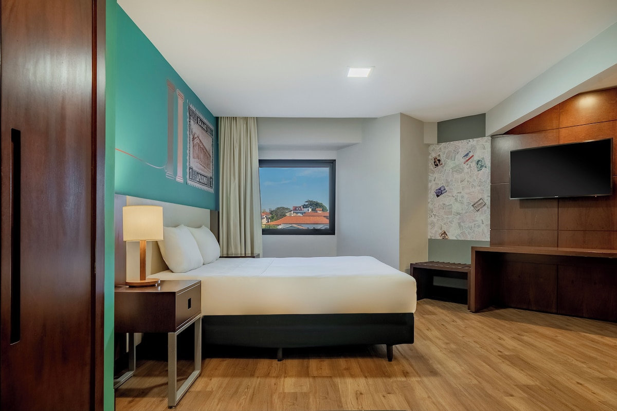 A hotel room with a bed, a nightstand, a lamp, a TV, and a window with a city view.