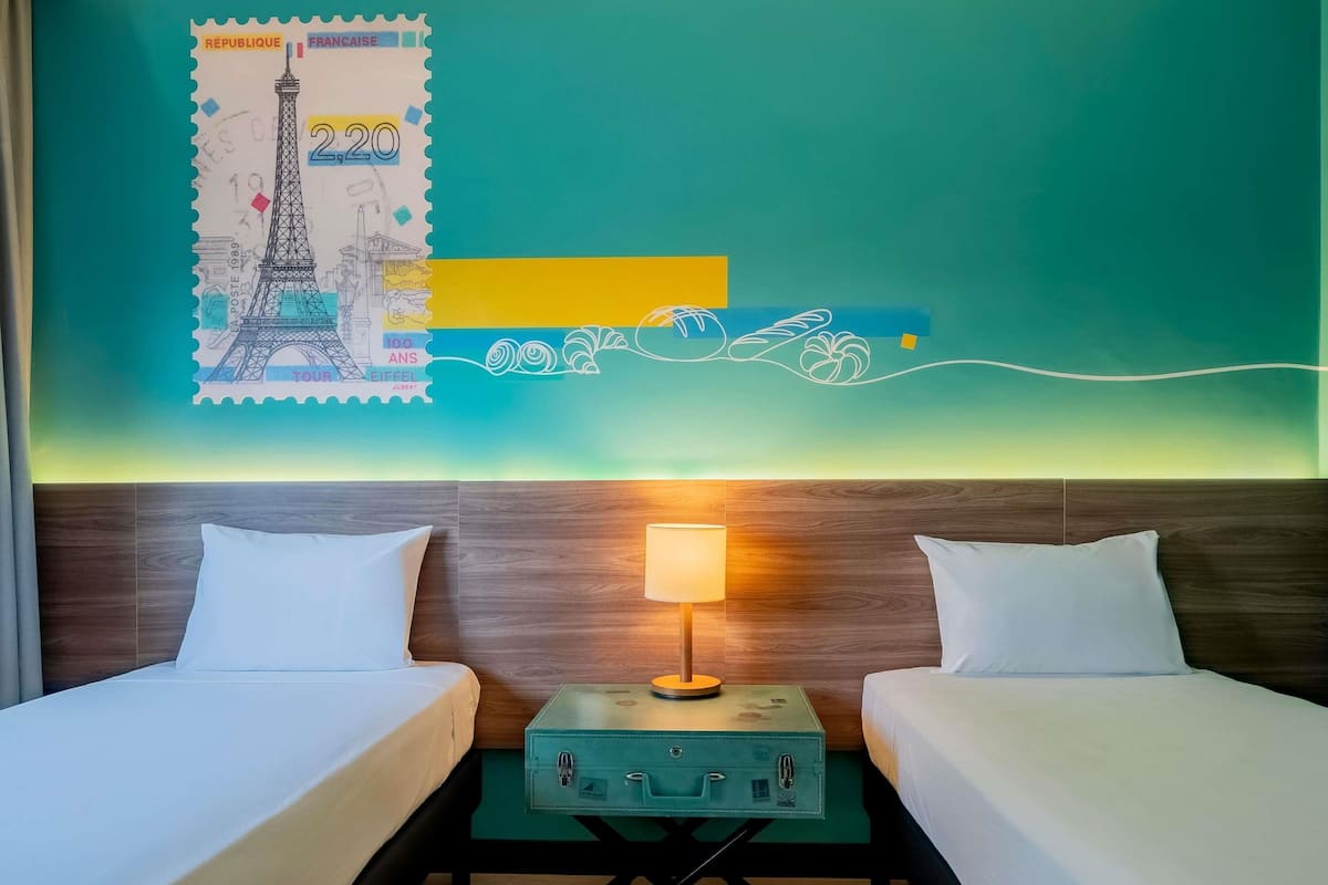 Une chambre d’hôtel avec deux lits, une table de chevet avec une lampe, une valise verte et un mur orné d’un timbre français et d’éléments décoratifs.