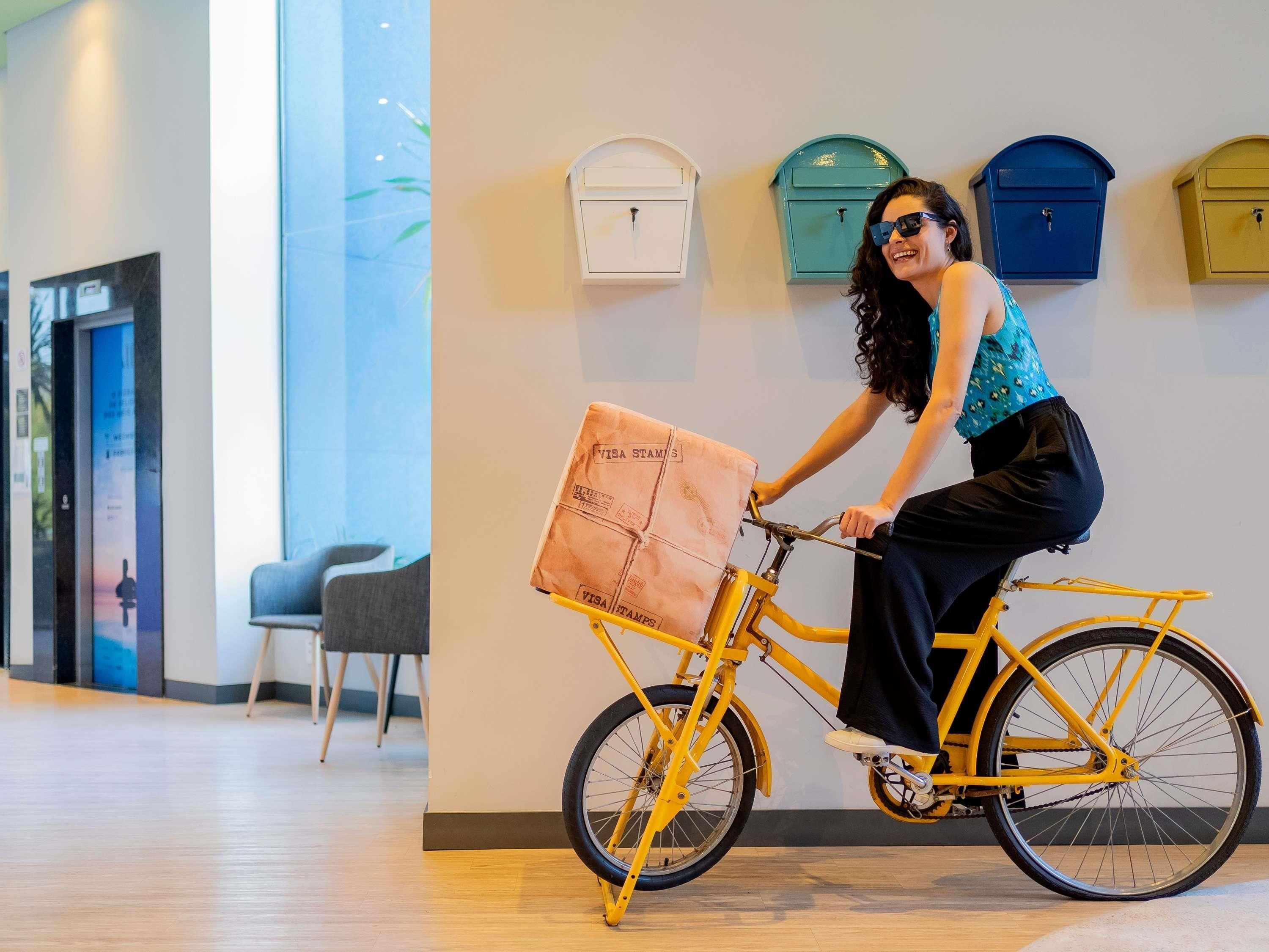 A person riding a yellow bicycle with a delivery box in a modern indoor setting with lockers and a chair.