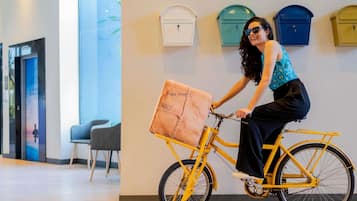 A person riding a yellow bicycle with a delivery box in a modern indoor setting with lockers and a chair.