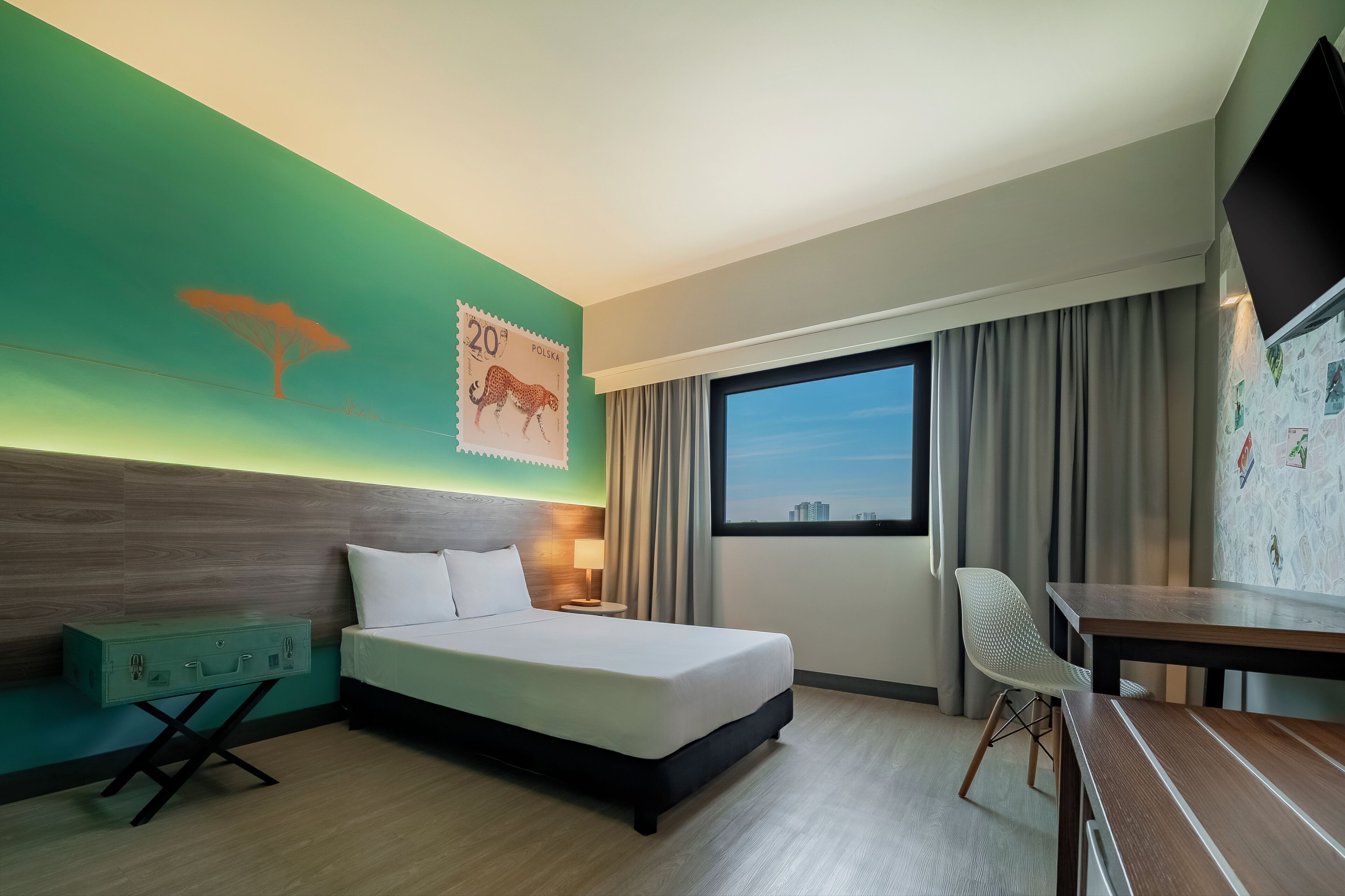 Habitación de hotel con cama, escritorio, silla, mesita de noche, lámpara, ventana con cortinas y un cartel en la pared.