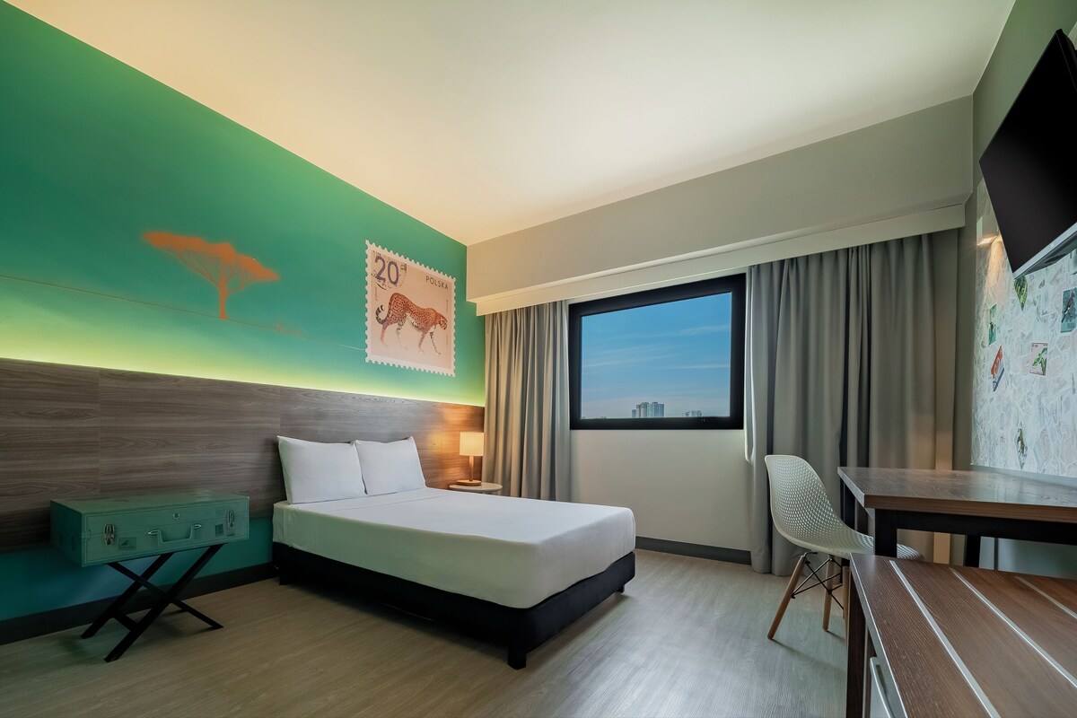 A hotel room with a bed, a desk, a chair, a nightstand, a lamp, a window with curtains, and a wall with a poster.