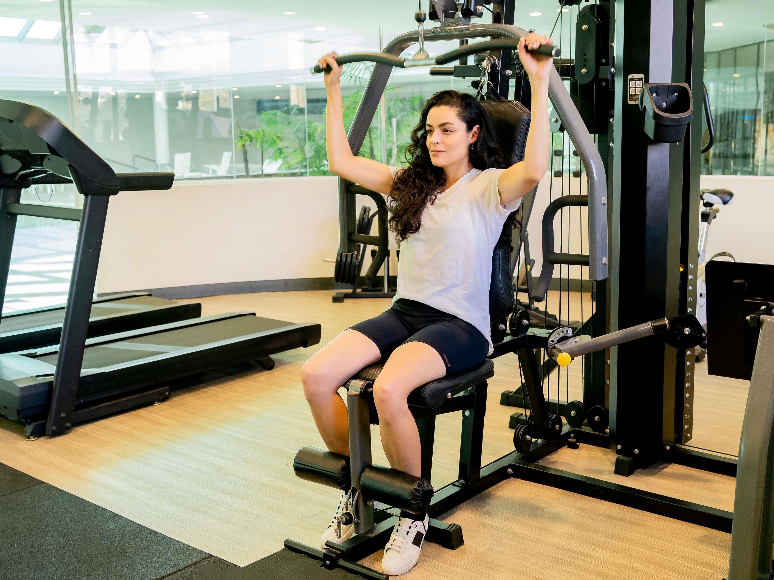 Una persona usando una máquina para piernas en un gimnasio, con una cinta de correr y pesas al fondo.