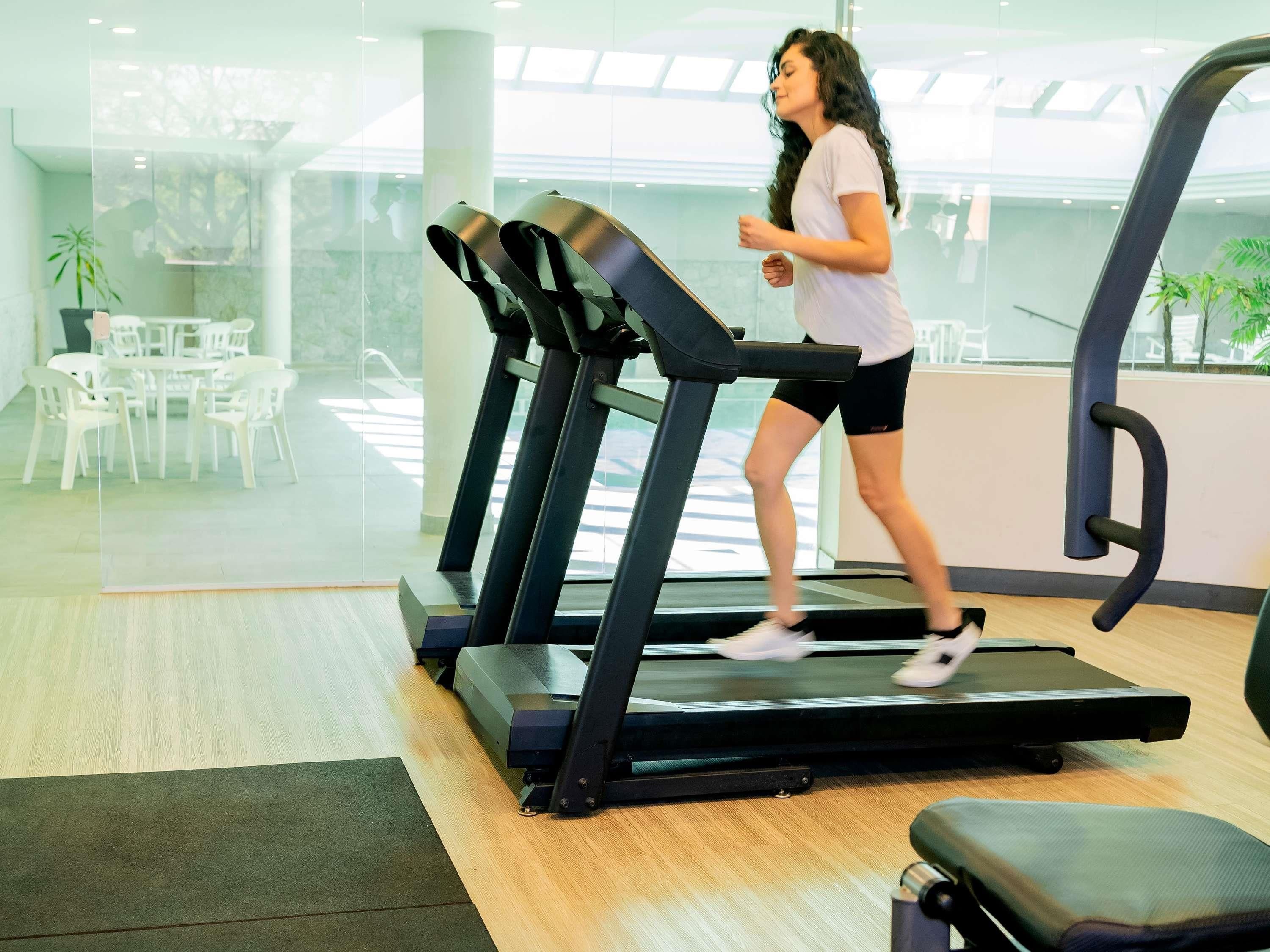 A woman running on a treadmill in a modern gym with glass walls, white chairs, and a wooden floor.