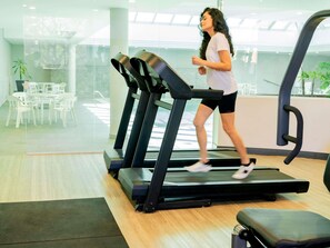 A woman running on a treadmill in a modern gym with glass walls, white chairs, and a wooden floor. - Ibis Styles Sorocaba Santa Rosalia (Sorocaba)