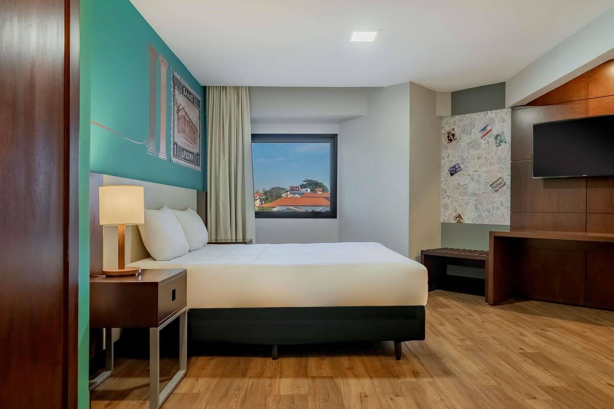 Une chambre d’hôtel avec un lit, une télévision, une fenêtre donnant sur un paysage agréable, un plancher en bois et un mur orné d’une affiche.