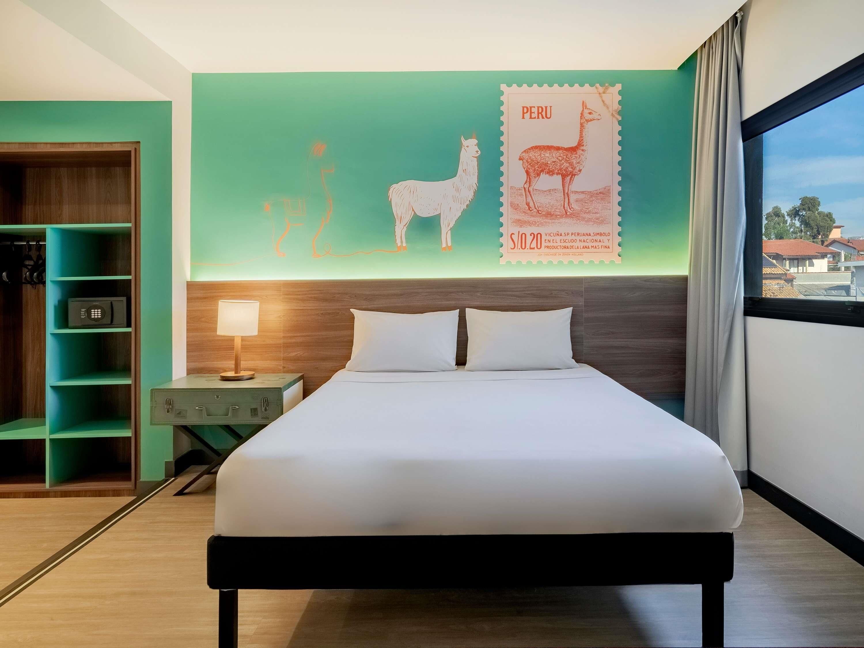 A hotel room with a bed, a nightstand, a lamp, a green wall with a Peruvian stamp, and a view of buildings through a window.