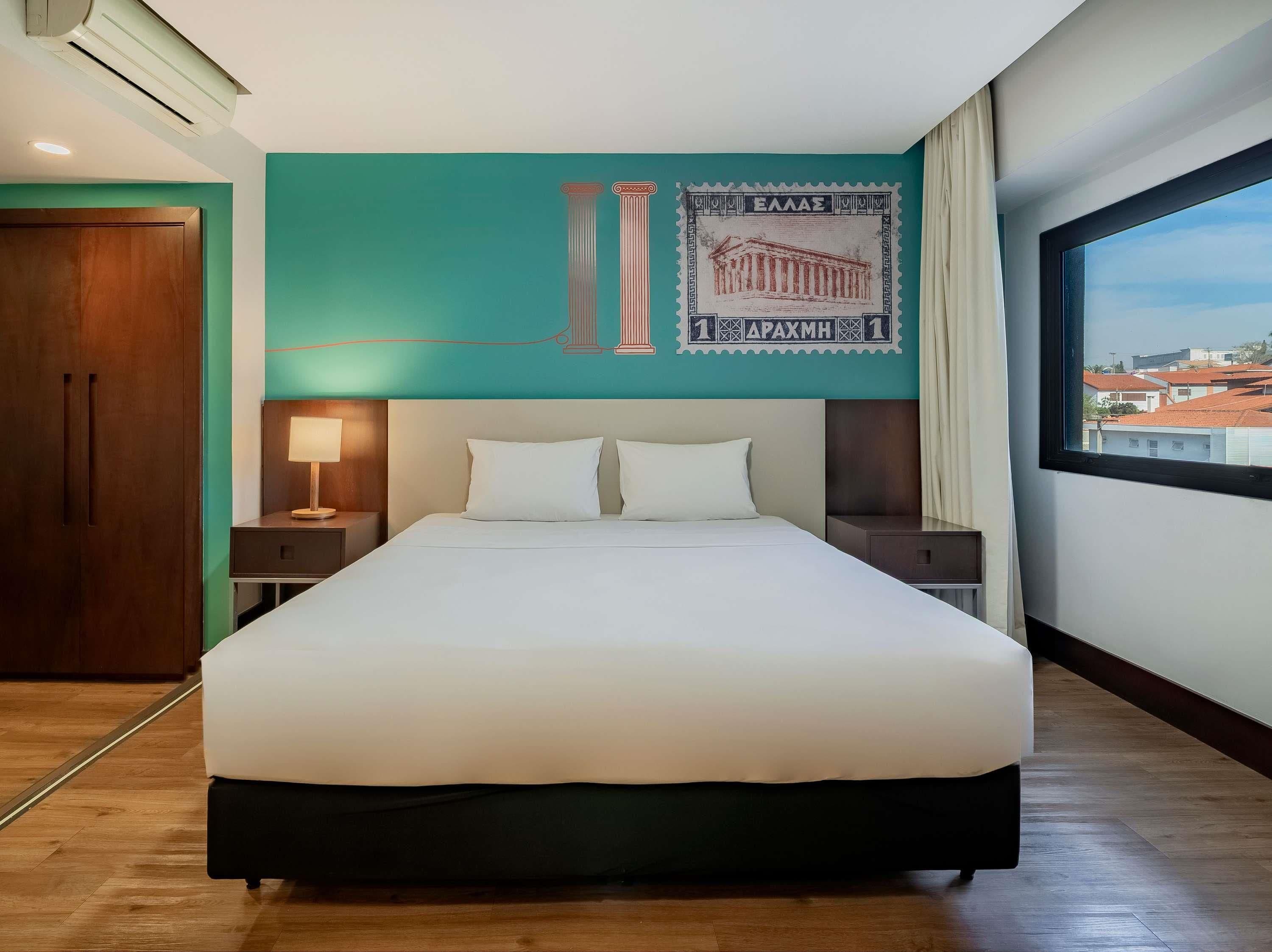 A hotel room with a large bed, two bedside tables, a wall-mounted air conditioner, and a window with a city view.