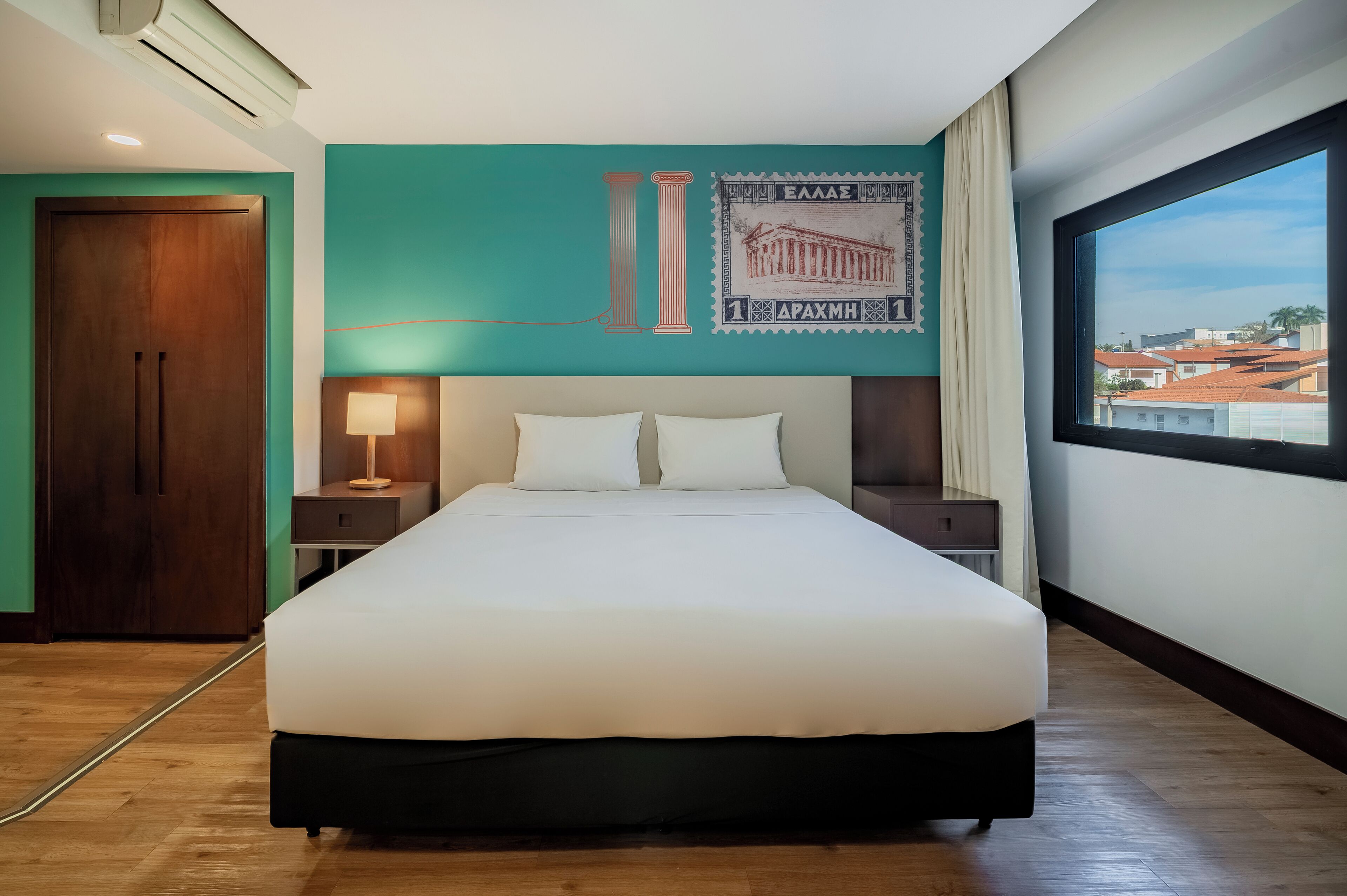Una habitación de hotel con una cama grande, una mesita de noche con una lámpara, una pared con un pilar y un sello, y una ventana con vistas a edificios.