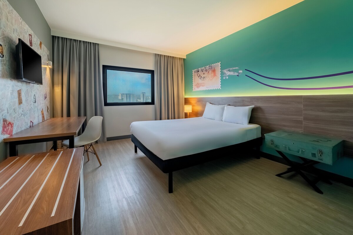 A hotel room with a bed, a desk, a chair, a television mounted on the wall, and a window with a city view.