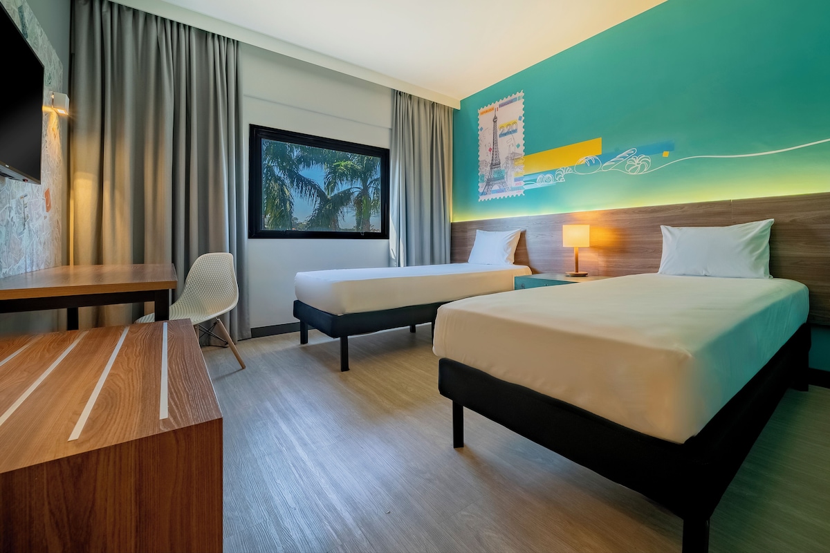 A hotel room with two beds, a desk, a chair, a TV, and a window with a view of palm trees.