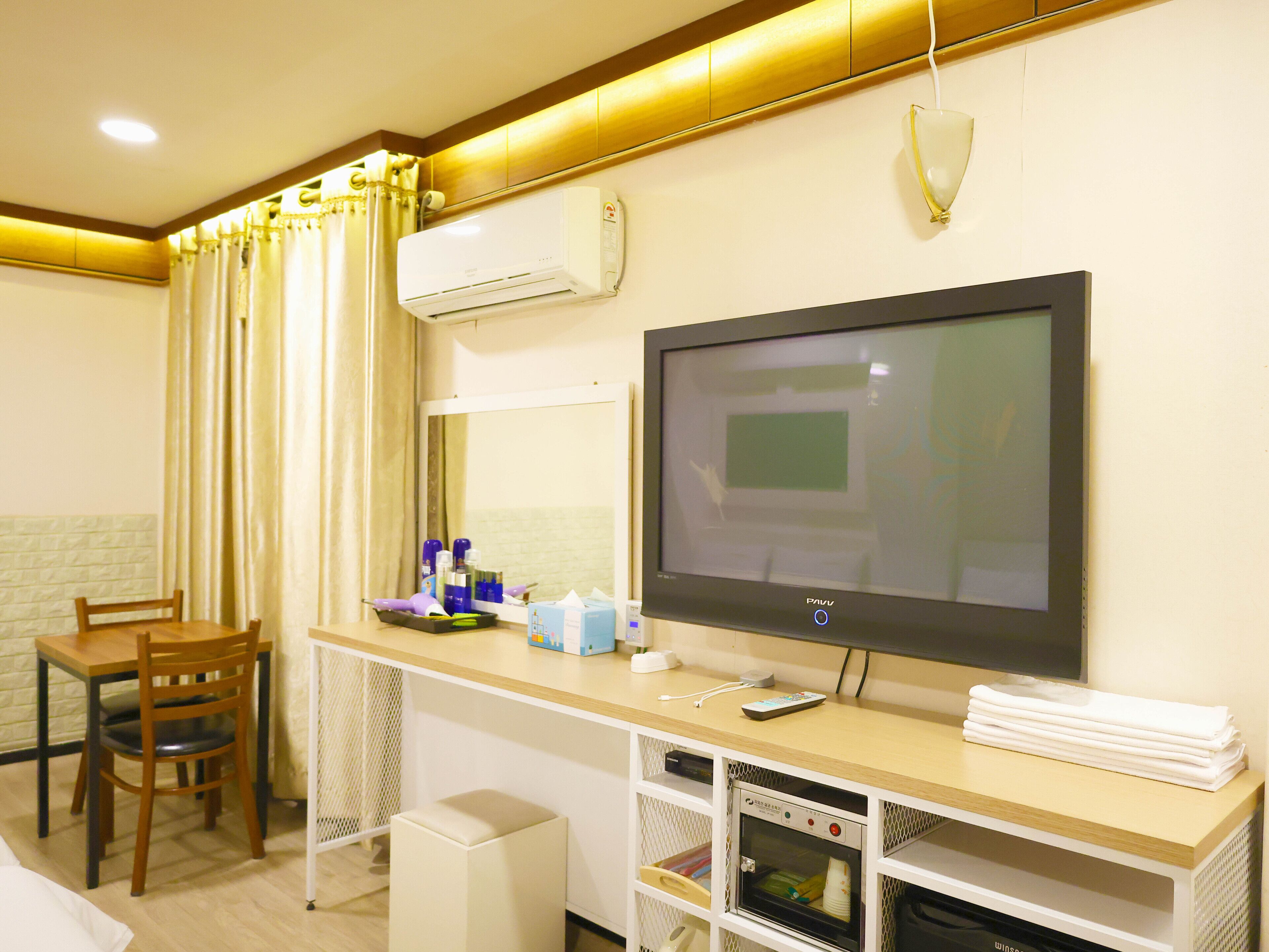 A hotel room with a TV, a desk with a computer, a chair, and a small kitchenette area.